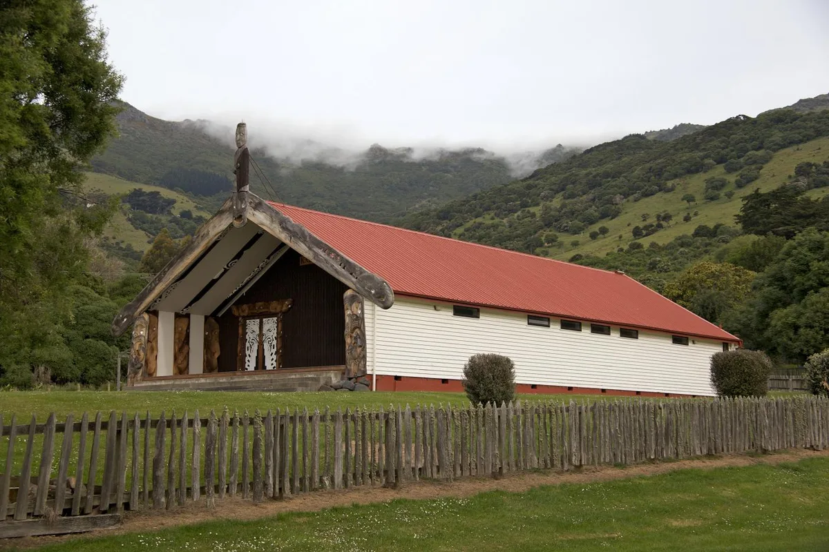 Ōnuku Marae: the carved wharenui with its red roof behind a picket fence, green hills of Akaroa behind.